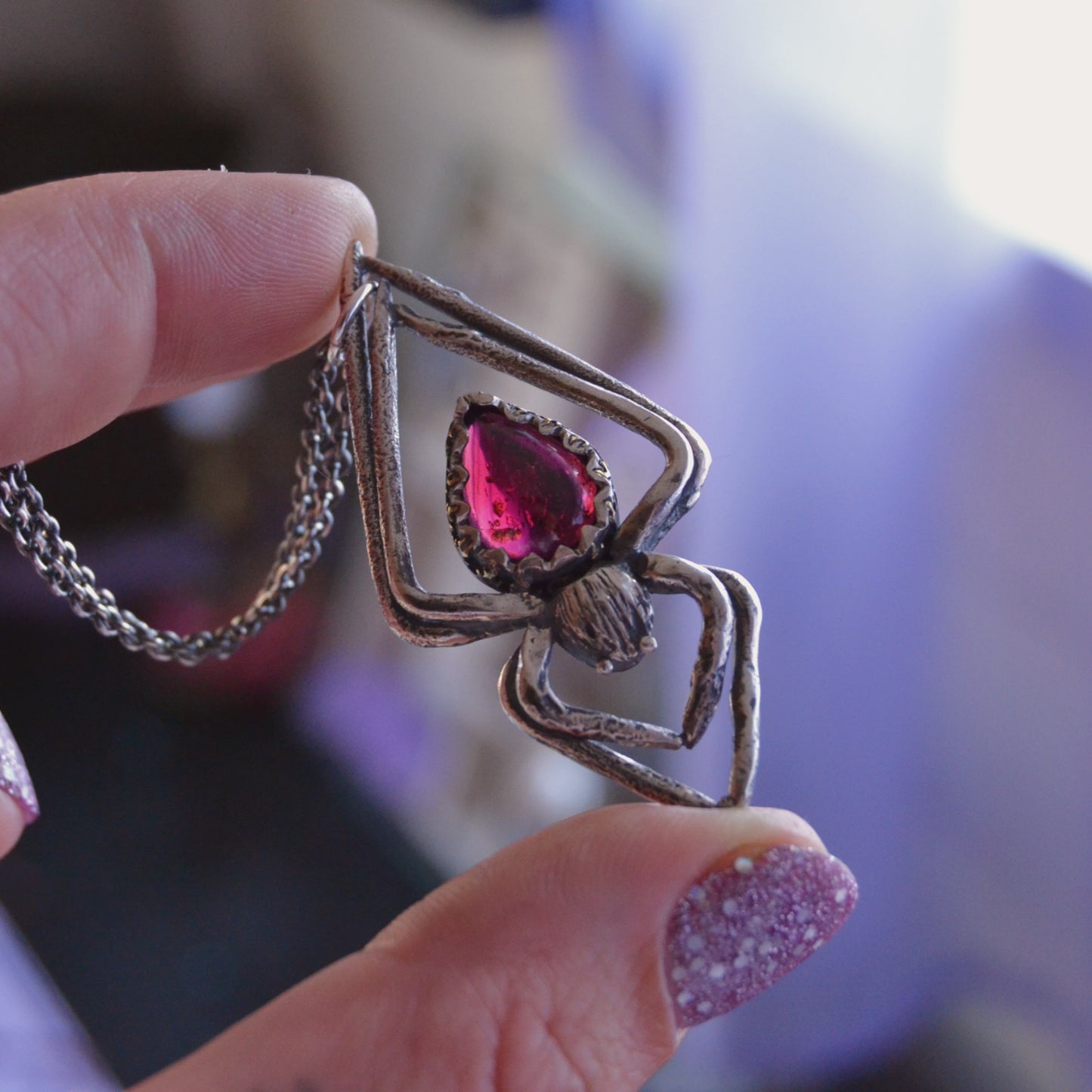 Handcrafted silver spider pendant necklace featuring a teardrop-shaped deep pink garnet gemstone set in a detailed, textured metal frame. The pendant is held between fingers against a softly blurred background.