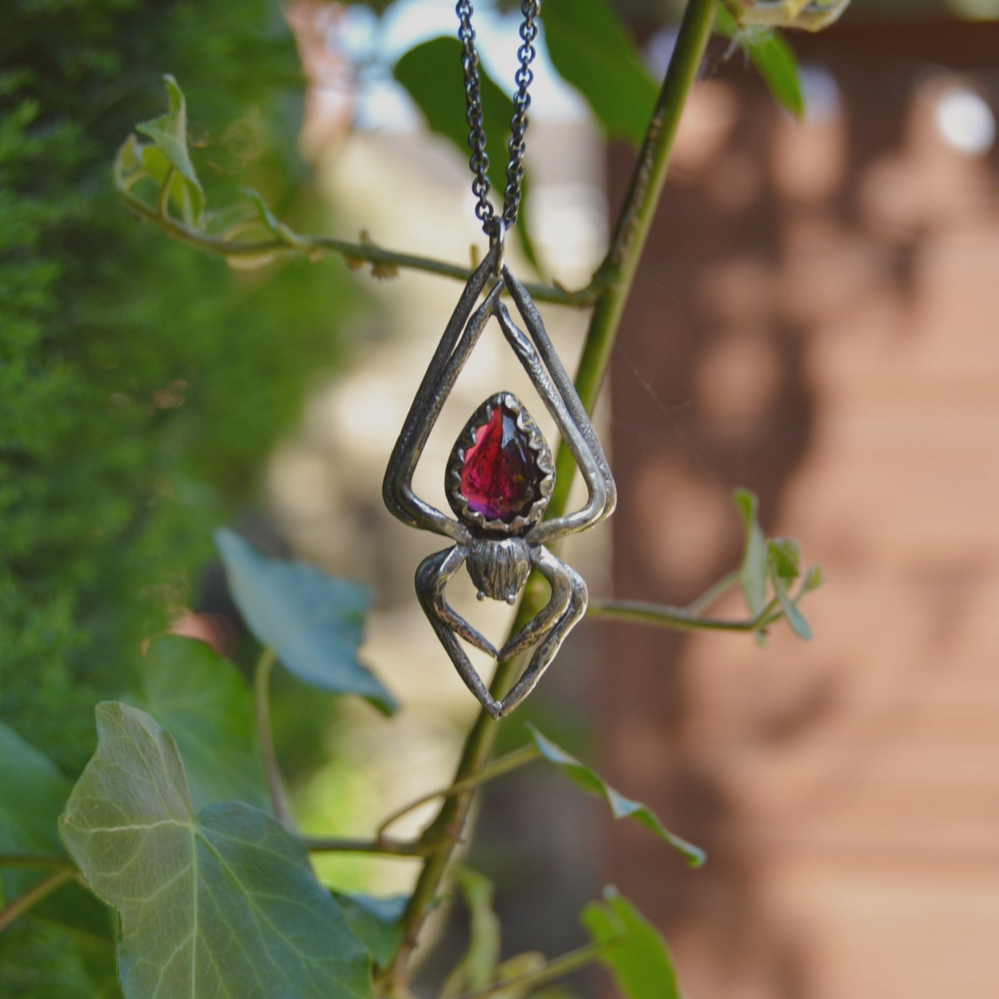 Handcrafted silver spider pendant necklace featuring a teardrop-shaped deep pink garnet gemstone set in a detailed, textured metal frame. The pendant is held between fingers against a softly blurred background.