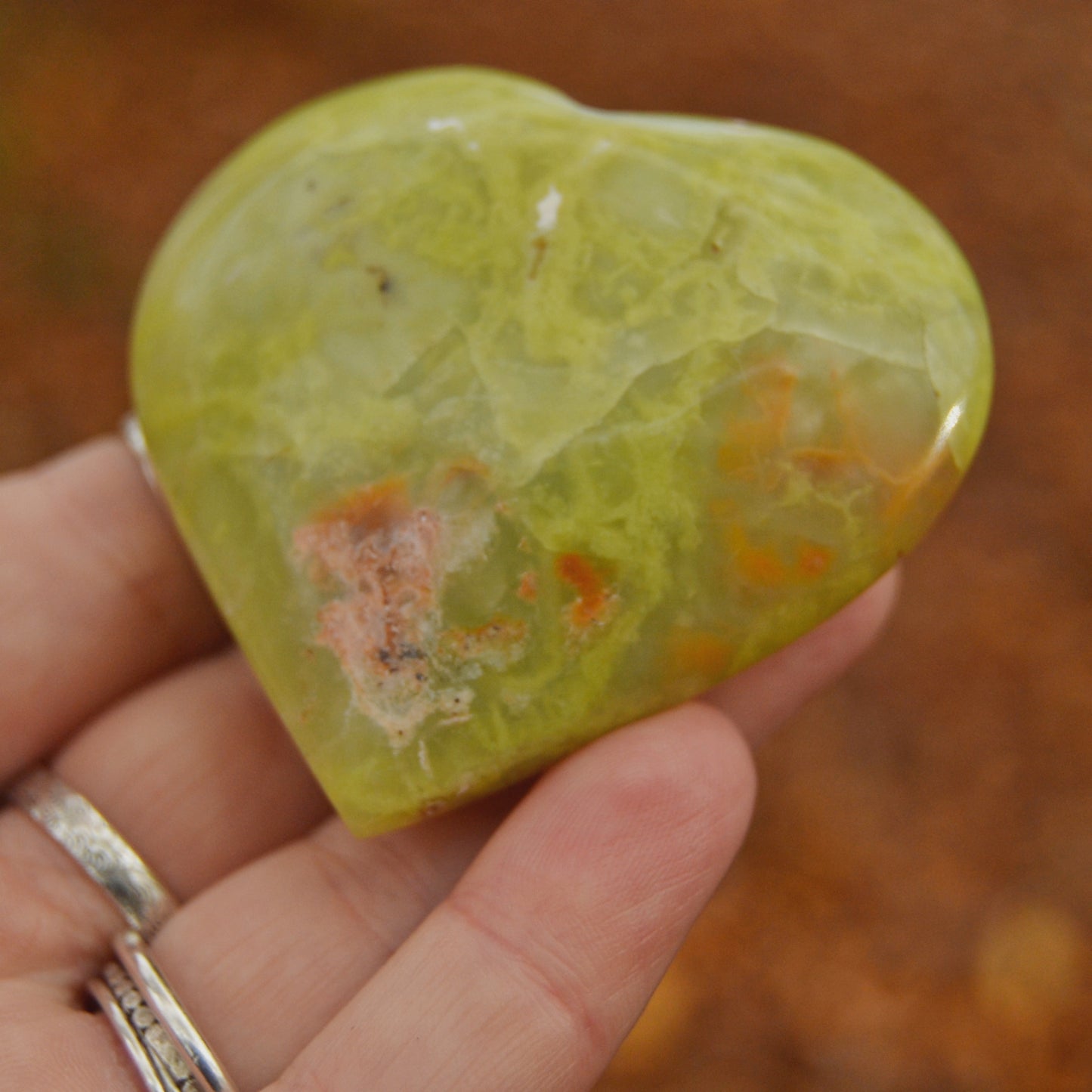 Hand holding a polished green serpentine crystal heart against an autumn forest background. The heart-shaped stone features natural marbling and earthy inclusions, with a glossy finish.