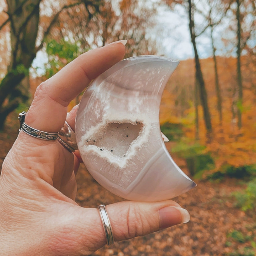 Polished agate crescent moon with a natural druzy geode centre, held in hand against a blurred autumnal forest background. The crystal features soft banding and sparkling druzy details.