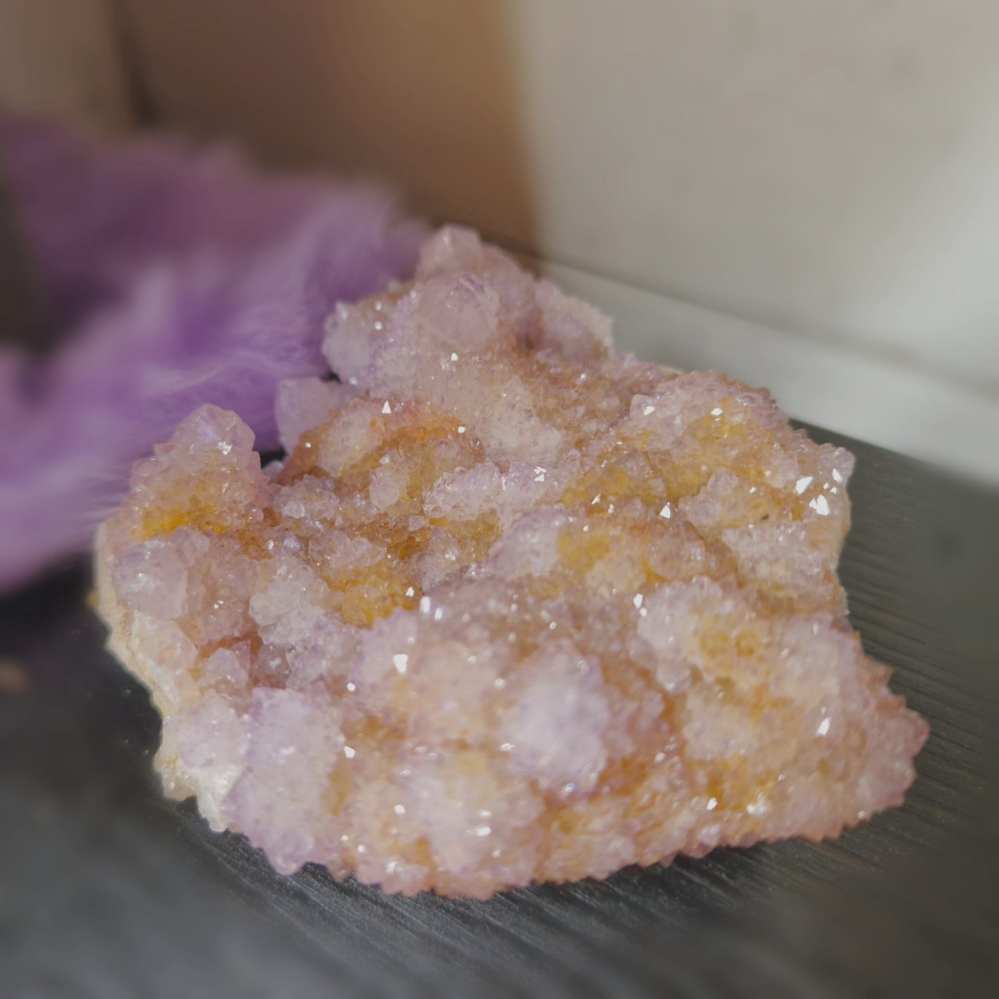 Close-up of a spirit quartz cluster with shimmering lavender and golden hues. The crystal features multiple small druzy points growing from a larger core, creating a sparkling, textured surface. Displayed on a dark surface with a soft purple background.