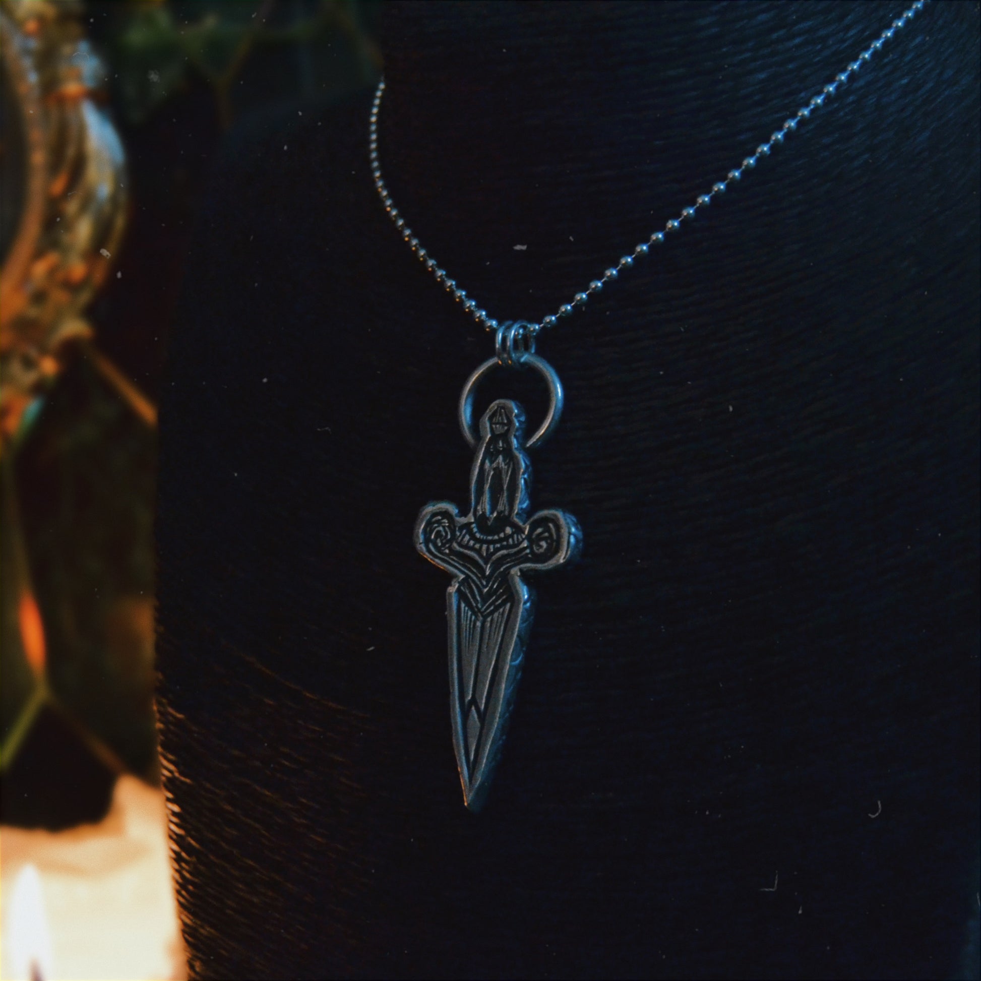 Silver dagger pendant necklace featuring intricate detailing and a figure of a woman at the top, displayed on a black bust stand. The background is dimly lit with autumnal decor, including a pumpkin.







