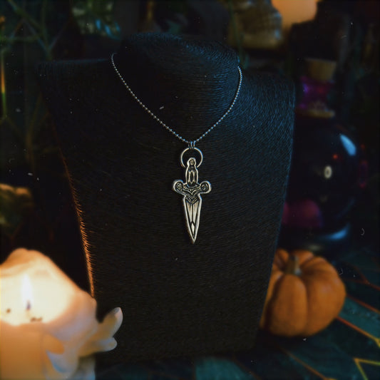 Silver dagger pendant necklace featuring intricate detailing and a figure of a woman at the top, displayed on a black bust stand. The background is dimly lit with autumnal decor, including a pumpkin.






