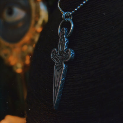 Silver dagger pendant necklace featuring intricate detailing and a figure of a woman at the top, displayed on a black bust stand. The background is dimly lit with autumnal decor, including a pumpkin.






