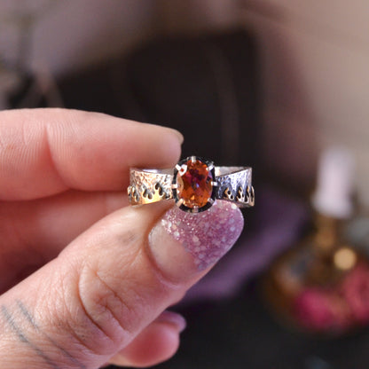 Close-up of a handcrafted sterling silver ring named "Like Fire," featuring a faceted round mystic topaz gemstone. The stone exhibits vibrant fiery hues of orange, red, and gold. The intricate crown-style setting holds the stone, and the wide silver band is adorned with organic flame-like patterns. The ring is worn on a tattooed finger, enhancing the artisan and bohemian aesthetic.

