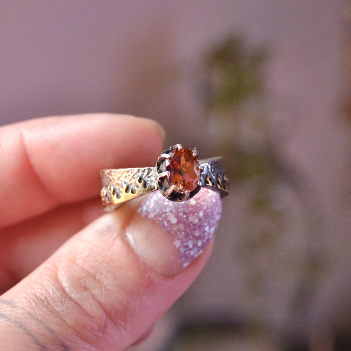 Close-up of a handcrafted sterling silver ring named "Like Fire," featuring a faceted round mystic topaz gemstone. The stone exhibits vibrant fiery hues of orange, red, and gold. The intricate crown-style setting holds the stone, and the wide silver band is adorned with organic flame-like patterns. The ring is worn on a tattooed finger, enhancing the artisan and bohemian aesthetic.

