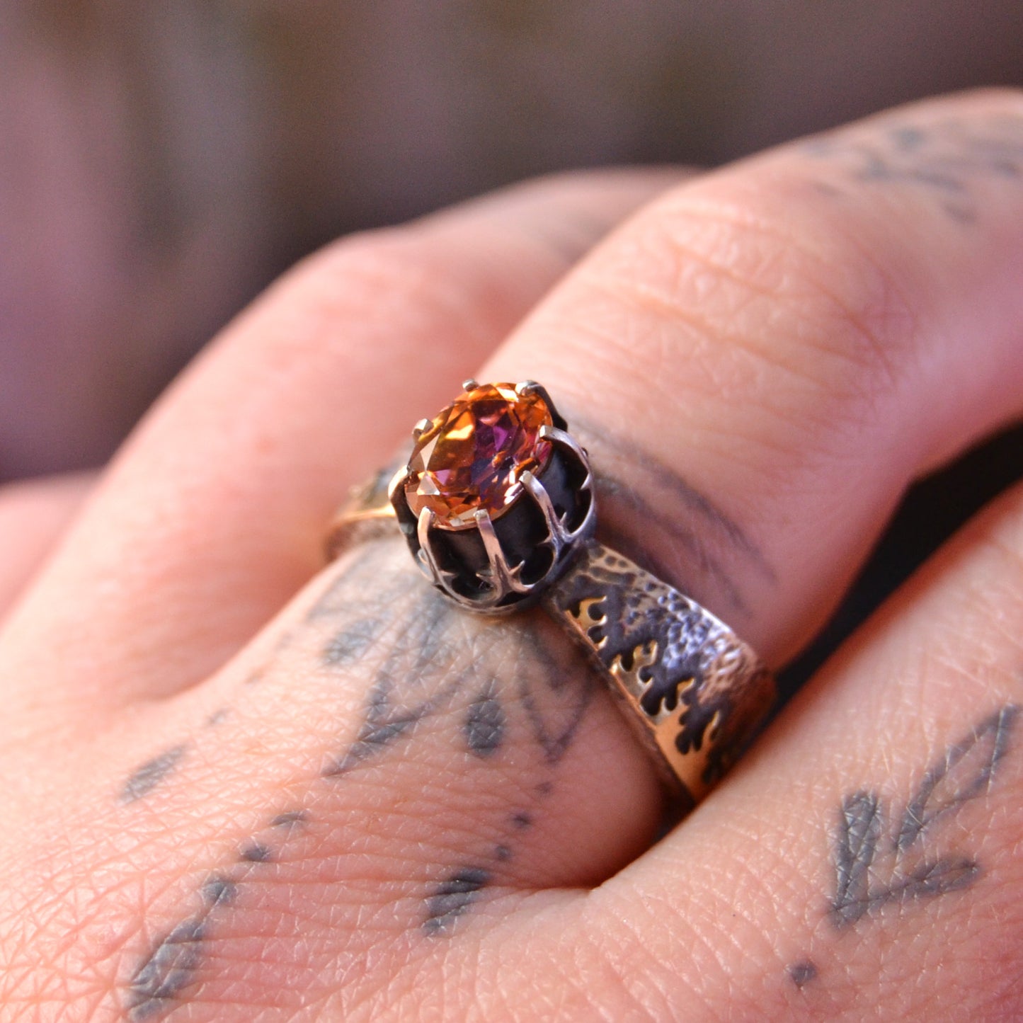 Close-up of a handcrafted sterling silver ring named "Like Fire," featuring a faceted round mystic topaz gemstone. The stone exhibits vibrant fiery hues of orange, red, and gold. The intricate crown-style setting holds the stone, and the wide silver band is adorned with organic flame-like patterns. The ring is worn on a tattooed finger, enhancing the artisan and bohemian aesthetic.

