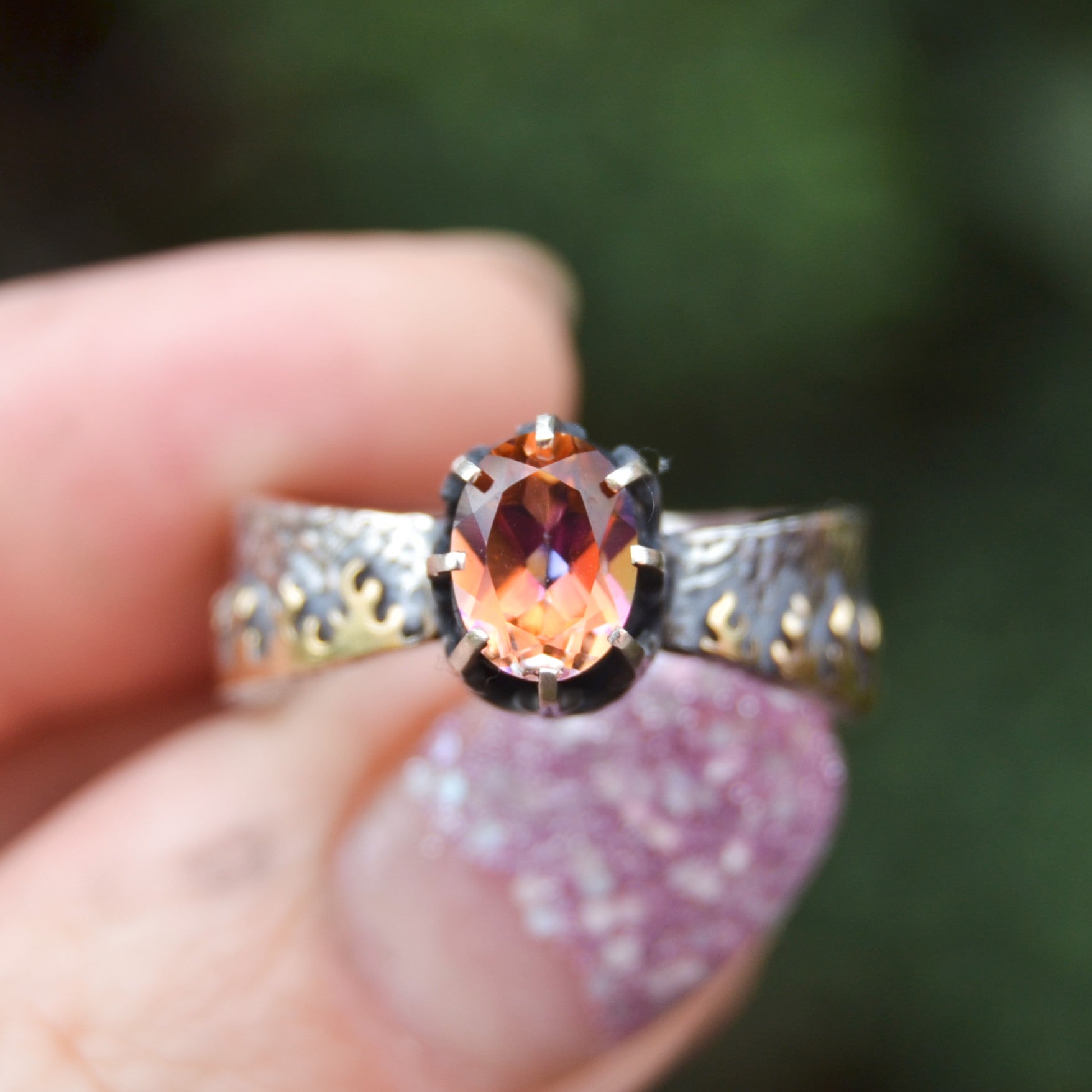 Close-up of a handcrafted sterling silver ring named "Like Fire," featuring a faceted round mystic topaz gemstone. The stone exhibits vibrant fiery hues of orange, red, and gold. The intricate crown-style setting holds the stone, and the wide silver band is adorned with organic flame-like patterns. The ring is worn on a tattooed finger, enhancing the artisan and bohemian aesthetic.

