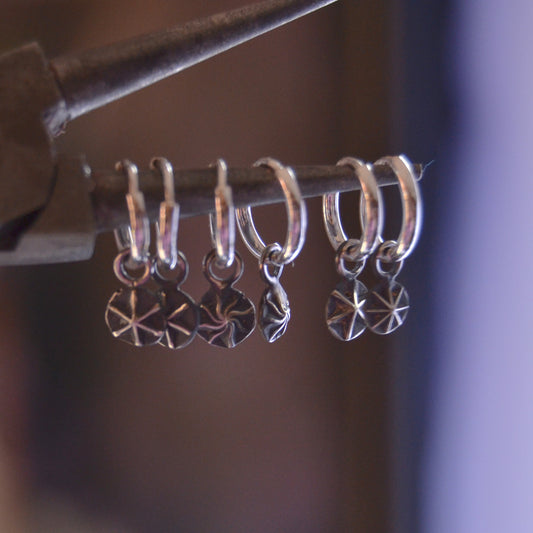 Close-up of handcrafted sterling silver hoop earrings with dangling star-etched charms. The earrings are displayed hanging on a rod, showcasing their polished finish and intricate celestial design.


