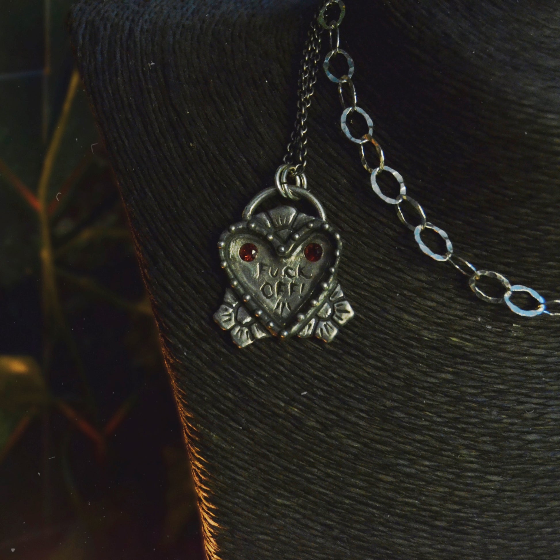 Close-up of a handcrafted sterling silver heart-shaped pendant with two bezel-set garnet stones. The pendant features the engraved words "F*** Off" and hangs from a mixed-link chain. The design is intricate, bold, and unique.

