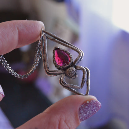 Handcrafted silver spider pendant necklace featuring a teardrop-shaped deep pink garnet gemstone set in a detailed, textured metal frame. The pendant is held between fingers against a softly blurred background.






