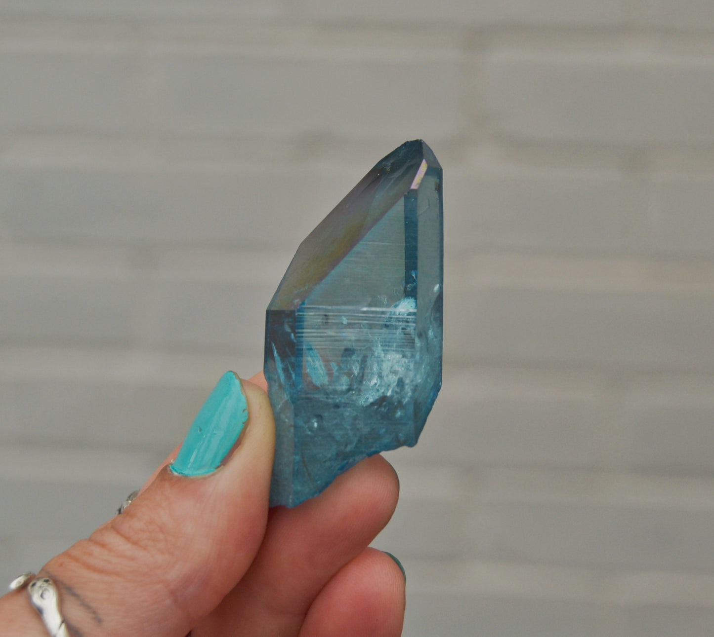 Vibrant blue polished quartz crystal point with natural inclusions and rough edges, held between fingers against a blurred outdoor background with a yellow flower and blue surface. The crystal’s surface is partially transparent with light reflecting off its facets.






