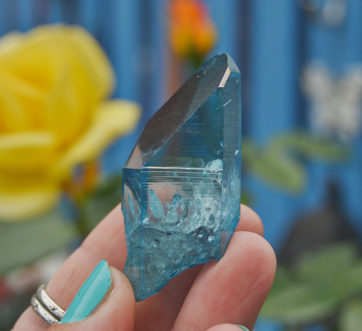 Vibrant blue polished quartz crystal point with natural inclusions and rough edges, held between fingers against a blurred outdoor background with a yellow flower and blue surface. The crystal’s surface is partially transparent with light reflecting off its facets.






