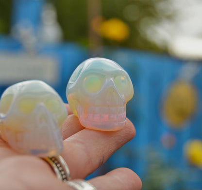 Small carved opalite crystal skull with an iridescent glow, held between fingers against a blurred outdoor background. The skull reflects hues of blue, pink, and yellow under natural light.







