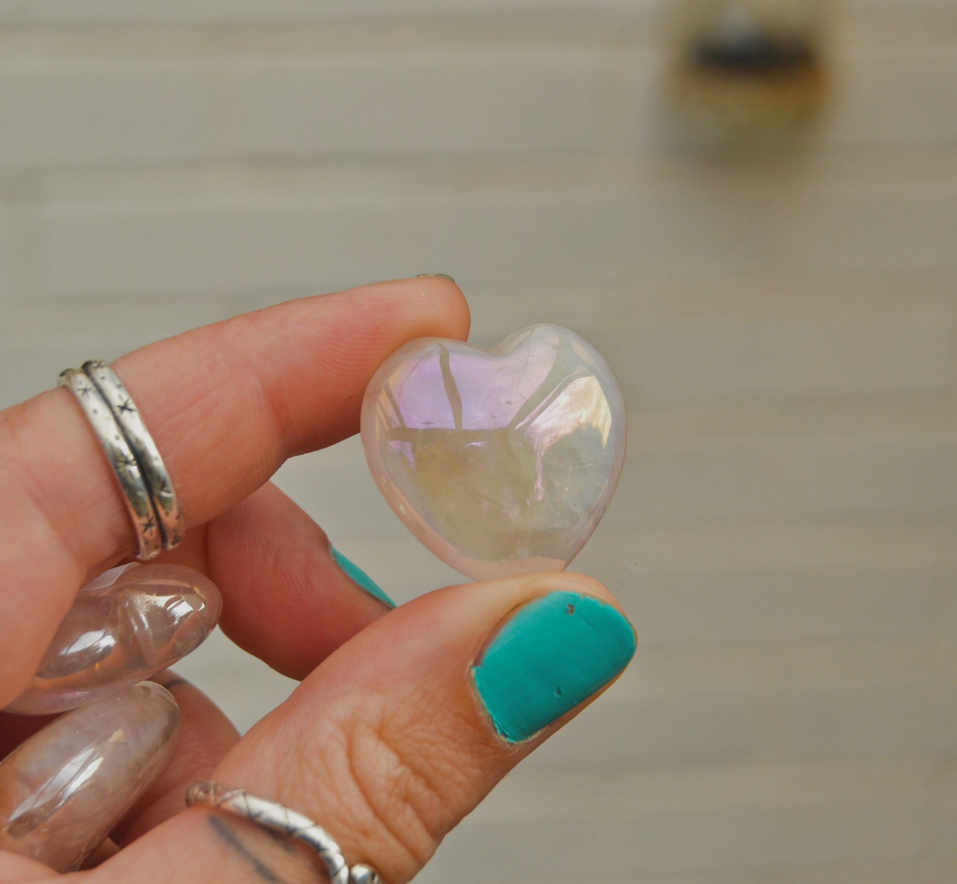 Small polished aura quartz heart with an iridescent sheen, held between fingers against a neutral background. The heart displays soft pastel reflections of pink, purple, and gold.






