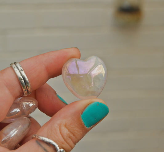 Small polished aura quartz heart with an iridescent sheen, held between fingers against a neutral background. The heart displays soft pastel reflections of pink, purple, and gold.






