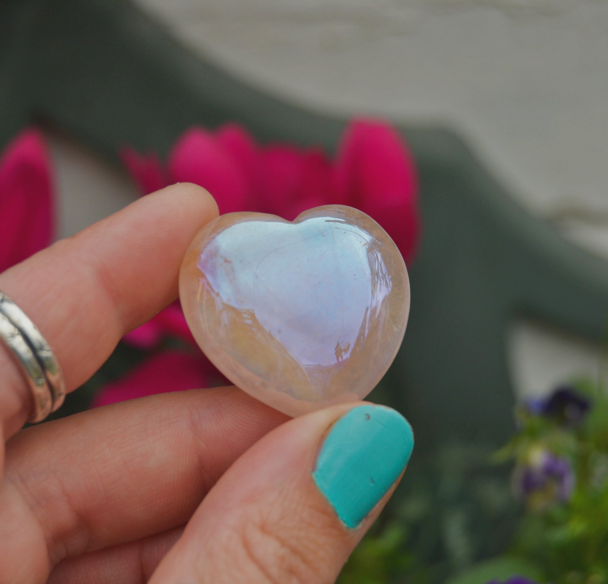 Small polished aura quartz heart with an iridescent sheen, held between fingers against a neutral background. The heart displays soft pastel reflections of pink, purple, and gold.






