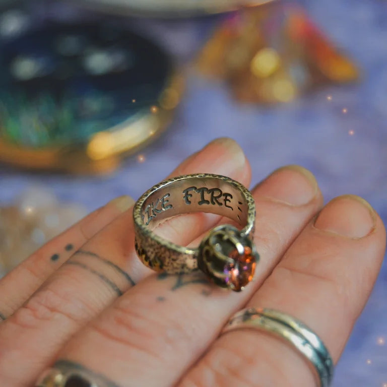 Close-up of a handcrafted sterling silver ring named "Like Fire," featuring a faceted round mystic topaz gemstone. The stone exhibits vibrant fiery hues of orange, red, and gold. The intricate crown-style setting holds the stone, and the wide silver band is adorned with organic flame-like patterns. The ring is worn on a tattooed finger, enhancing the artisan and bohemian aesthetic.

