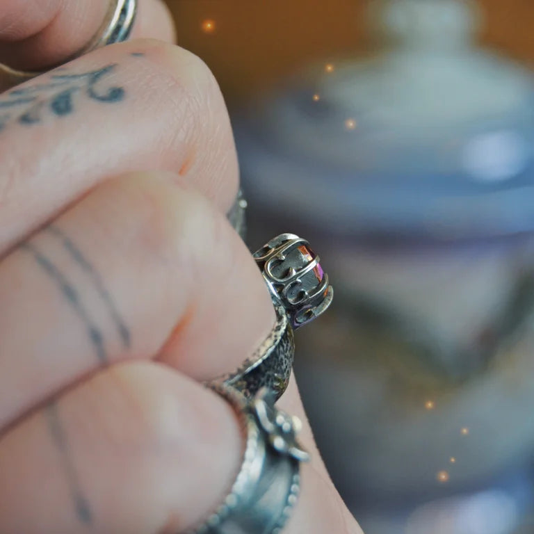 Close-up of a handcrafted sterling silver ring named "Like Fire," featuring a faceted round mystic topaz gemstone. The stone exhibits vibrant fiery hues of orange, red, and gold. The intricate crown-style setting holds the stone, and the wide silver band is adorned with organic flame-like patterns. The ring is worn on a tattooed finger, enhancing the artisan and bohemian aesthetic.

